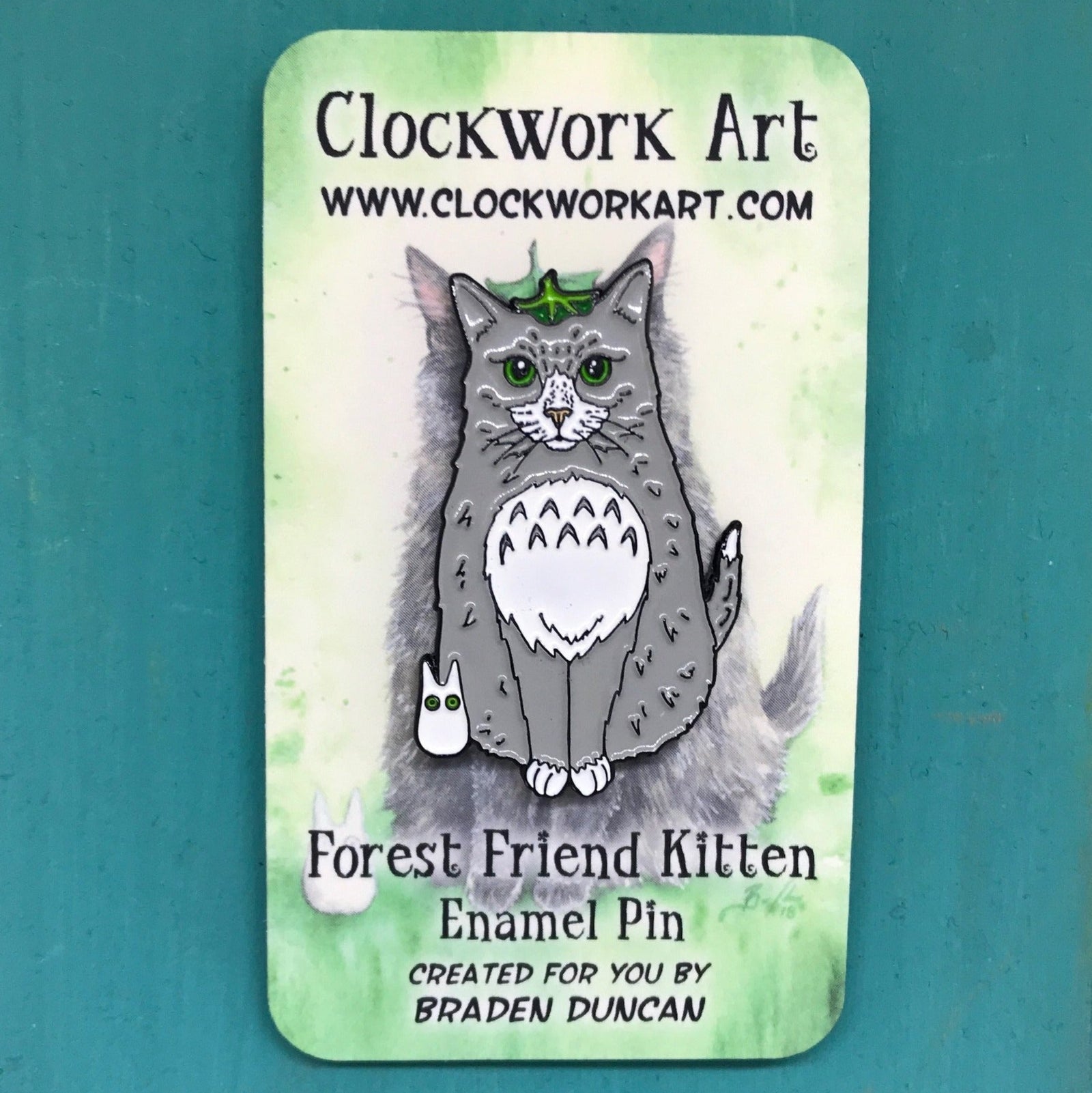 Enamel pin of a cat with a white belly on a green and white card with 'Clockwork Art' branding.