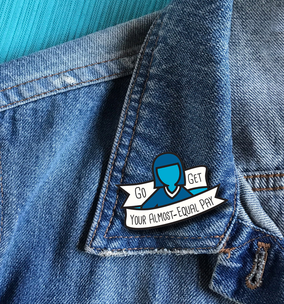 A close-up of a blue enamel pin attached to the collar of a denim jacket, with a turquoise ribbed shirt visible underneath. The pin features a stylized blue figure and white banners with the text 'Go GET YOUR ALMOST-EQUAL PAY'.