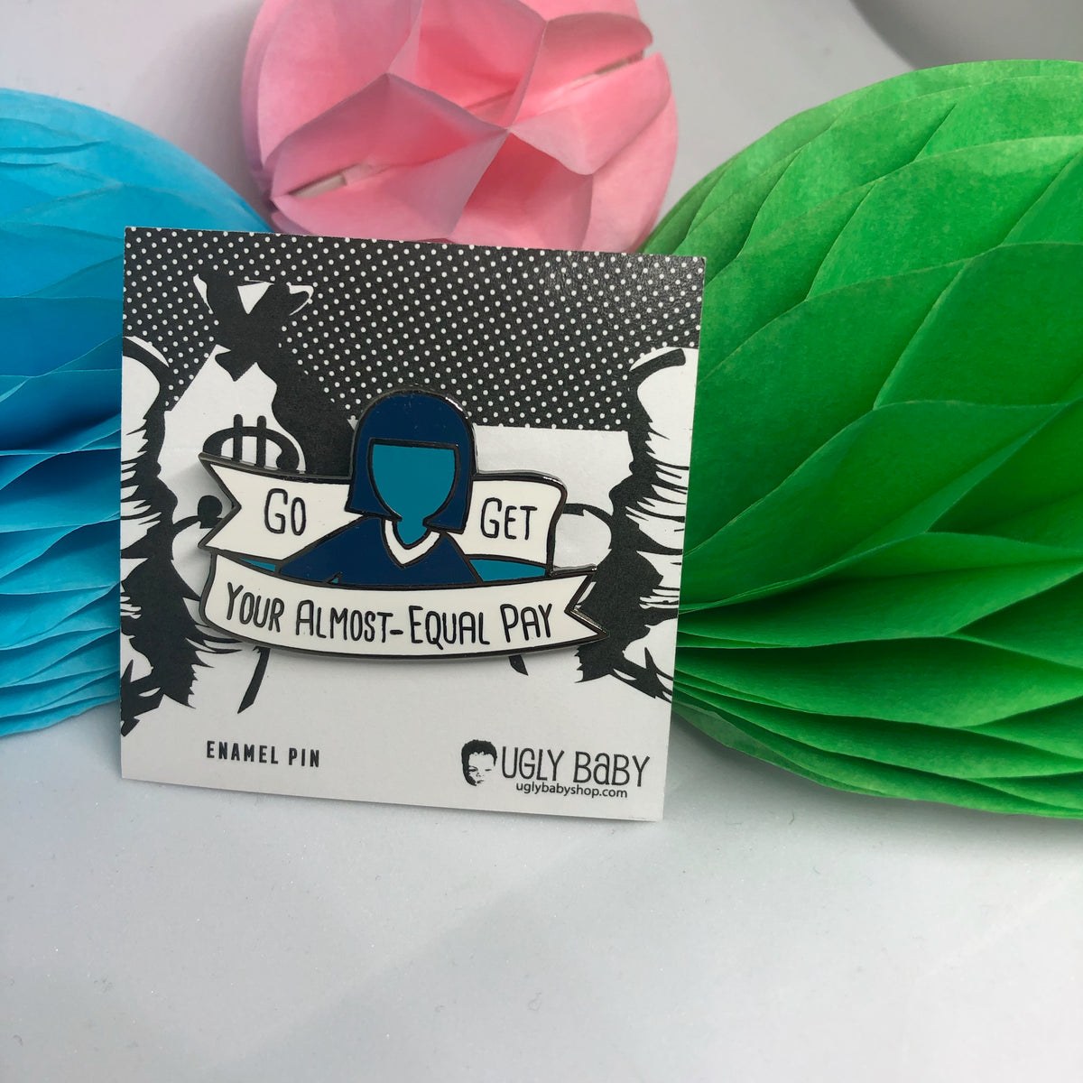 An enamel pin on its backing card, displayed in front of colorful paper honeycomb decorations. The pin features a stylized illustration of a person with a blue silhouette and black hair, holding a banner that reads 'Go GET Your ALMOST-EQUAL PAY'.