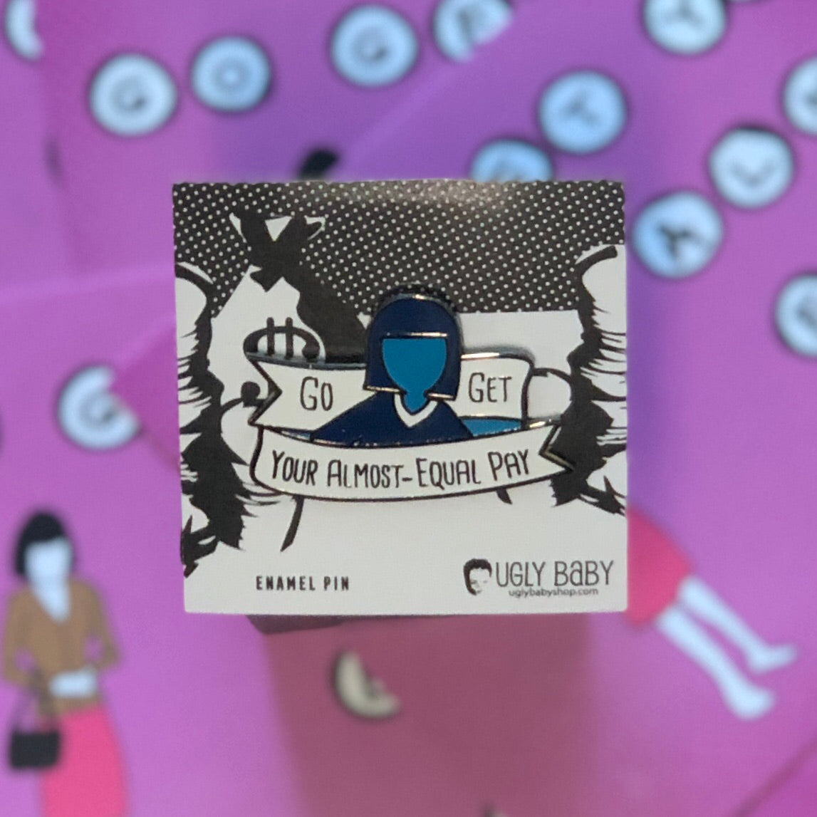 An enamel pin by Ugly Baby featuring a stylized blue and black illustration of a person holding a banner that reads 'Go GET YOUR ALMOST-EQUAL PAY', displayed on its white backing card against a pink background with cartoon graphics.