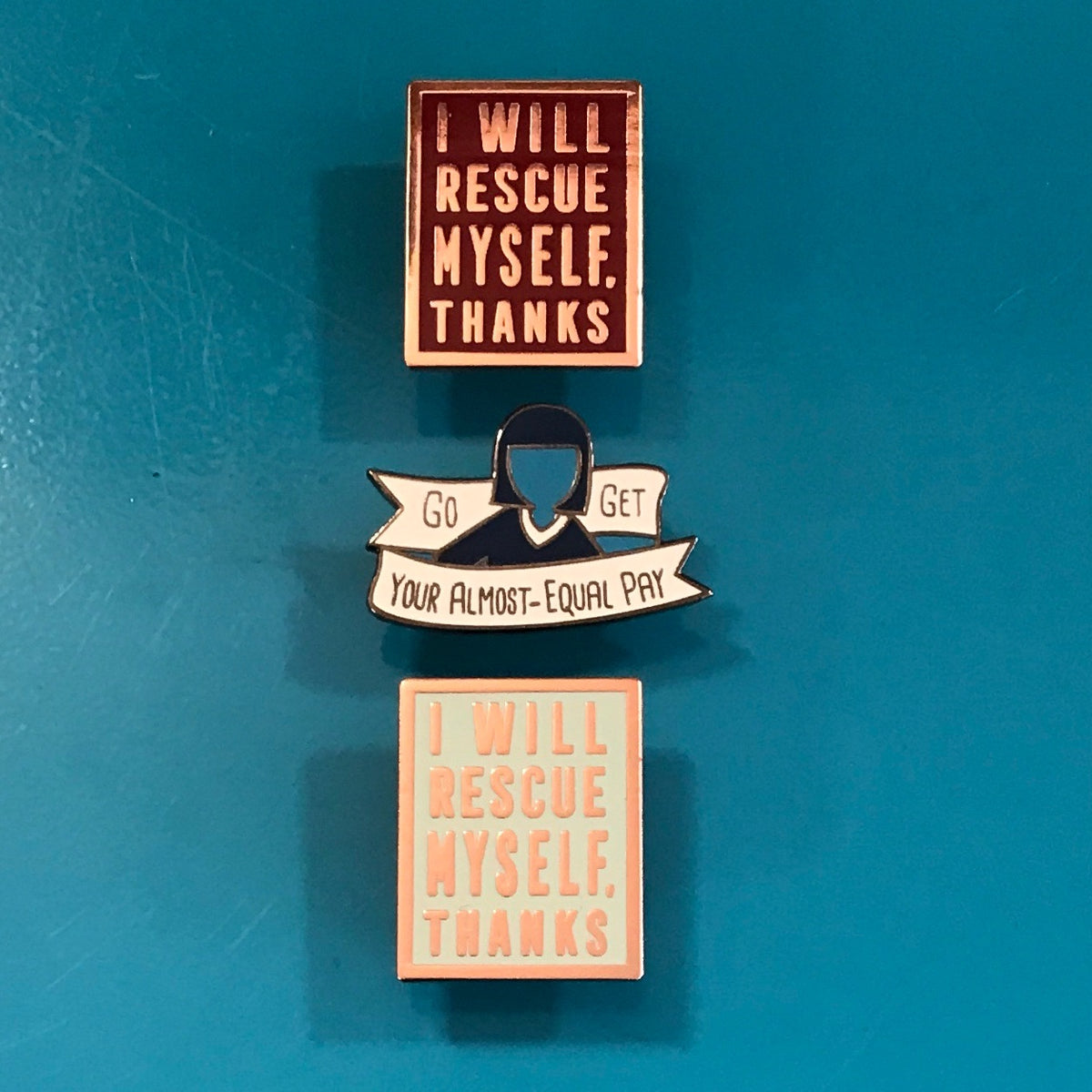 Three enamel pins with self-care and workplace advocacy messages are arranged vertically on a textured teal-blue background. The top pin is dark red with cream text reading 'I WILL RESCUE MYSELF.