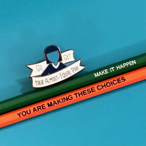 An enamel pin featuring a silhouette of a person with a blue hair bun and a white banner that reads 'Go GET YOUR ALMOST-EQUAL PAY', placed on top of two pencils, one green and one orange, on a solid blue background.