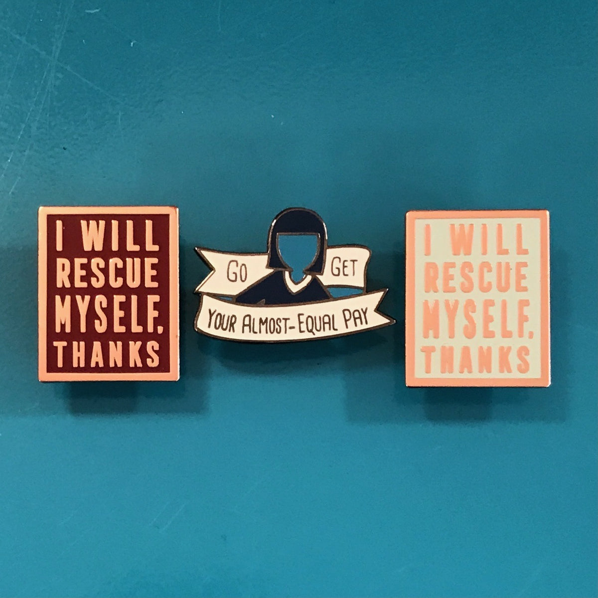 Three enamel pins displayed on a teal background. The left pin is square with a dark red background and rose gold text reading 'I WILL RESCUE MYSELF.