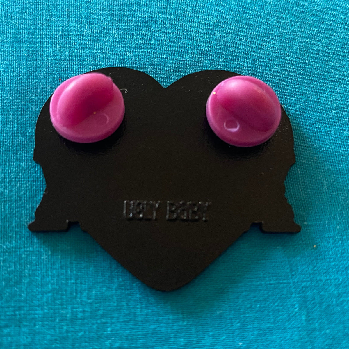 Back of a Heart shaped enamel pin with two pink rubber stoppers. "Ugly Baby" is stamped onto the back. 