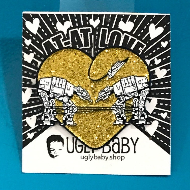 Heart shaped enamel pin called Love AT-AT First Sight with a gold glitter background. Shown on a backing card with hearts. 