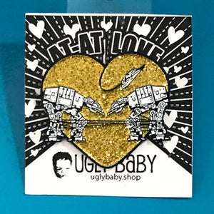 Heart shaped enamel pin called Love AT-AT First Sight with a gold glitter background. Shown on a backing card with hearts. 