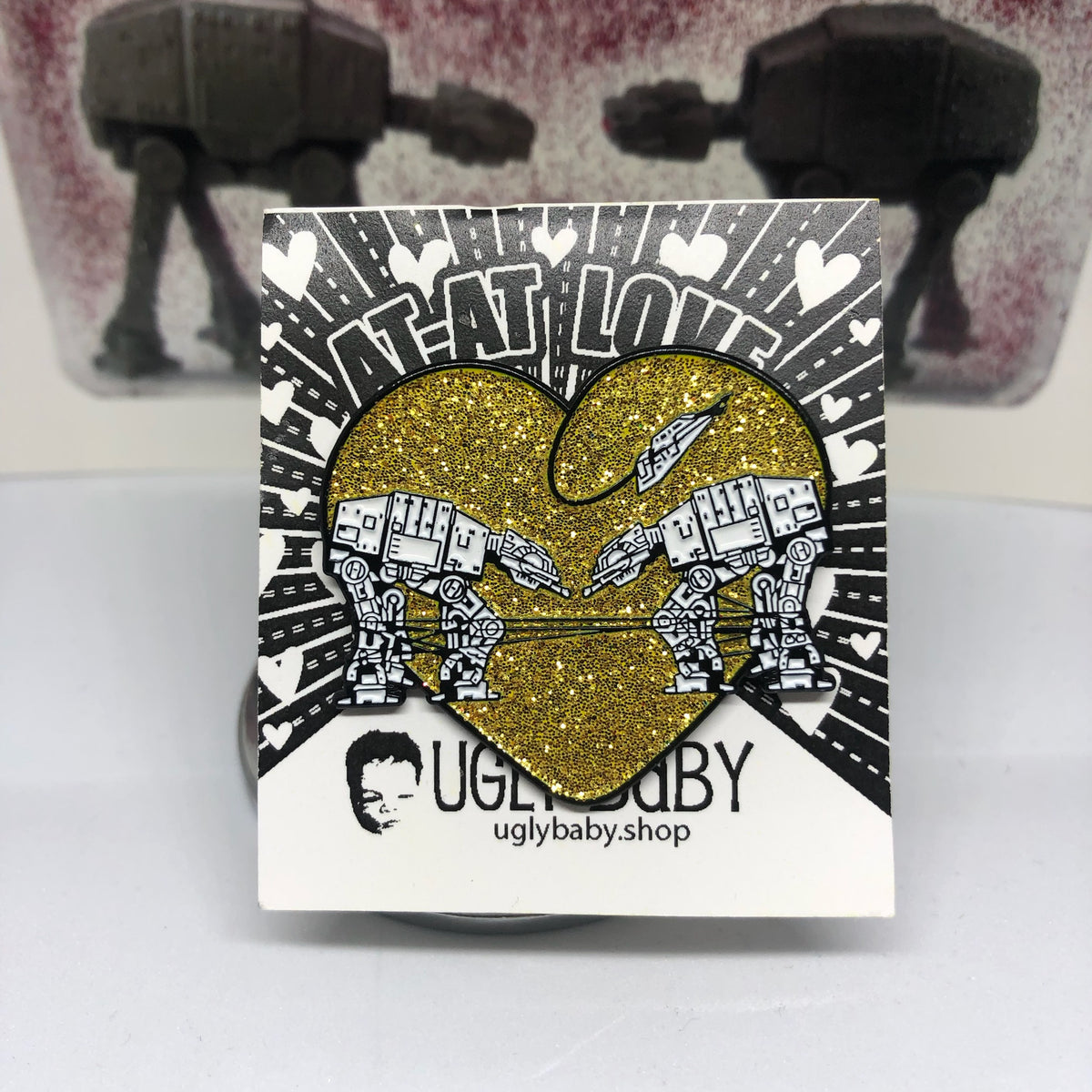 Heart shaped enamel pin called Love AT-AT First Sight with a gold glitter background. The original Shower Art piece that this design is based on is shown in the background. 