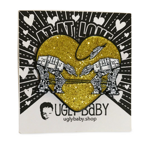Heart shaped enamel pin called Love AT-AT First Sight with a gold glitter background. Shown on a backing card with hearts. 