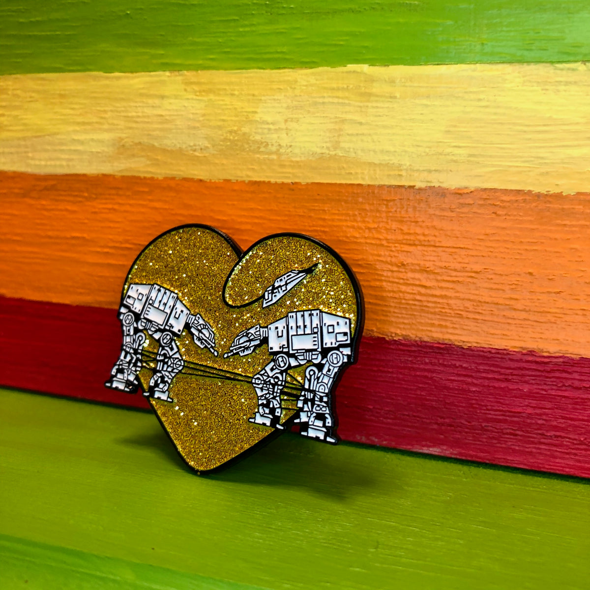 Heart shaped enamel pin called Love AT-AT First Sight with a gold glitter background. Shown against a rainbow background.