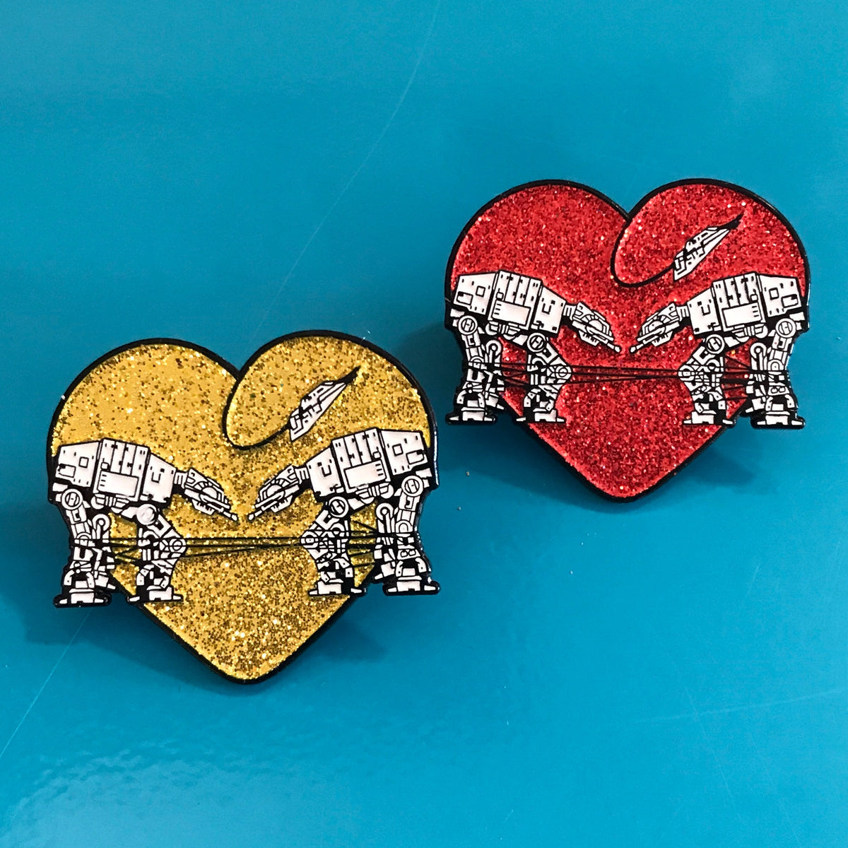 Heart shaped enamel pin called Love AT-AT First Sight with a gold glitter background. Shown with a matching enamel pin in red glitter. 