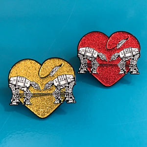 Heart shaped enamel pin called Love AT-AT First Sight with a gold glitter background. Shown with a matching enamel pin in red glitter. 