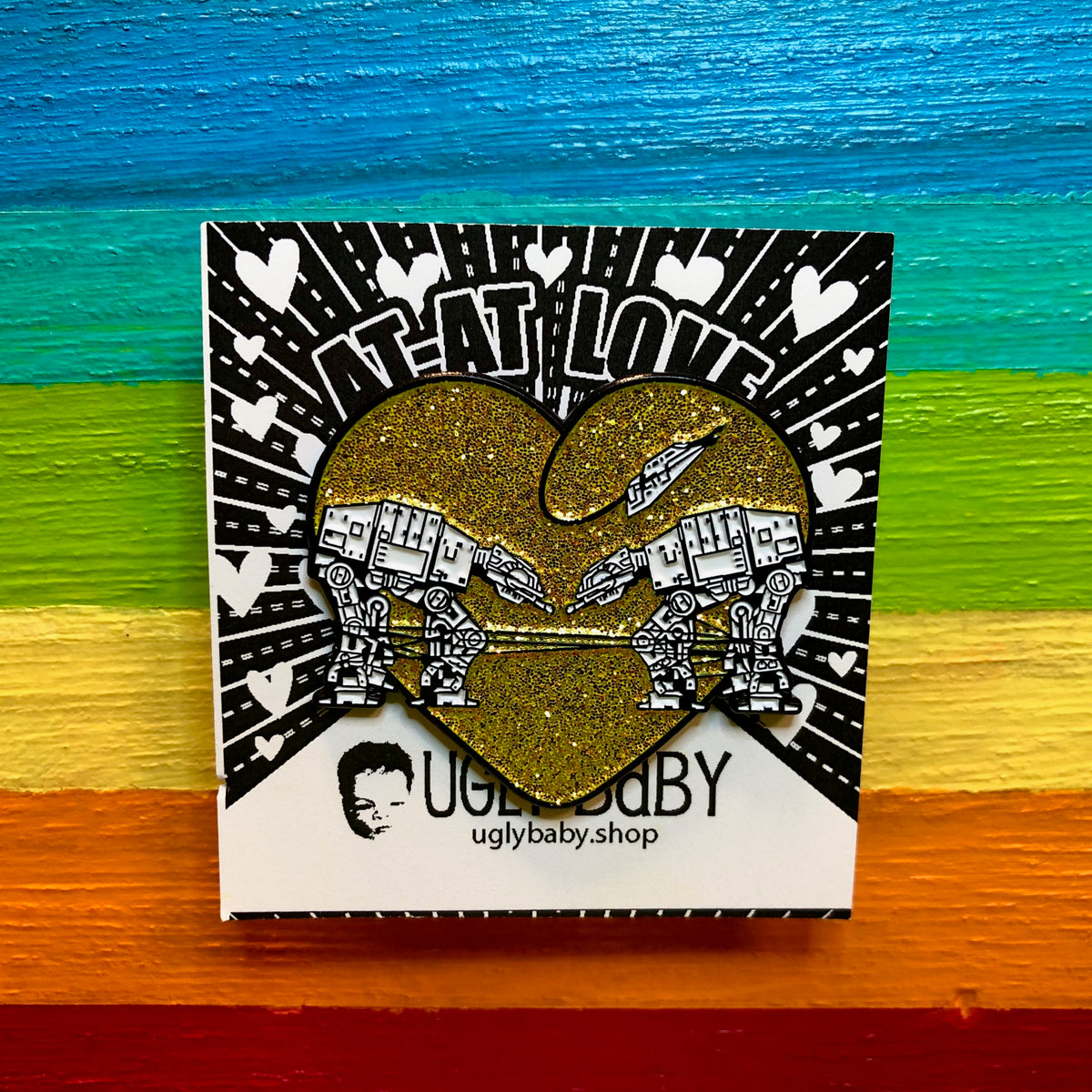 A square card featuring a glittery gold heart-shaped pin with AT-AT walkers against a rainbow-striped wooden background.