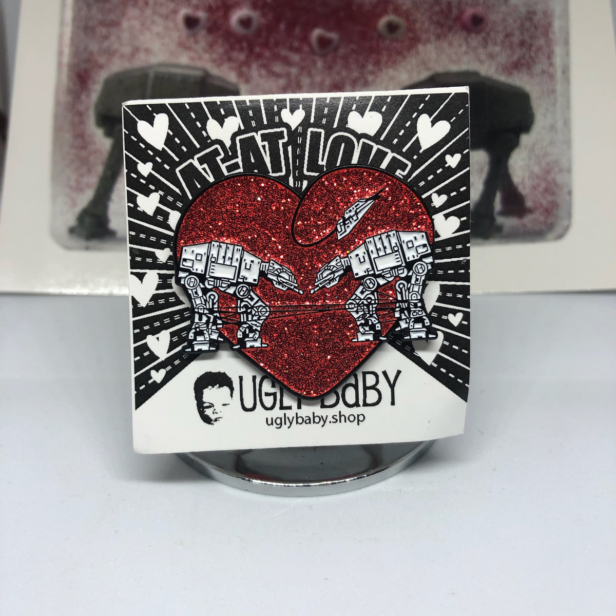 Heart shaped enamel pin called Love AT-AT First Sight with a red glitter background. Shown on a square paper background with black and white hearts. 