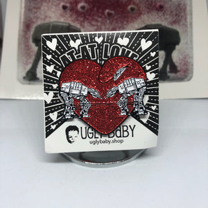 Heart shaped enamel pin called Love AT-AT First Sight with a red glitter background. Shown on a square paper background with black and white hearts. 