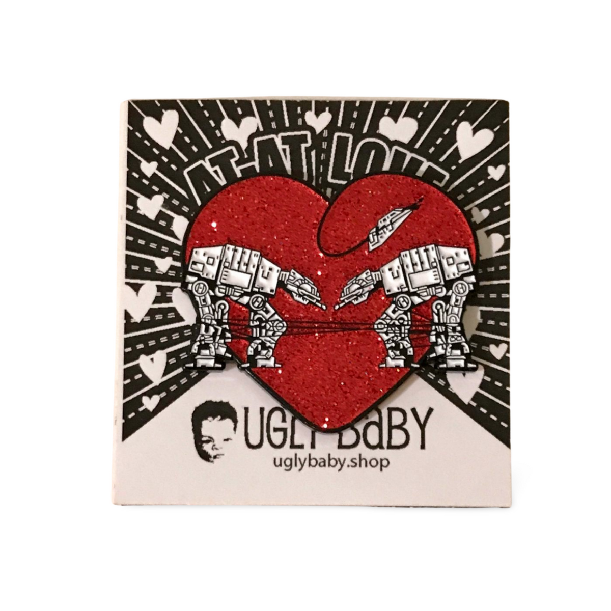Heart shaped enamel pin called Love AT-AT First Sight with a red glitter background. Shown on a square paper background with black and white hearts. 