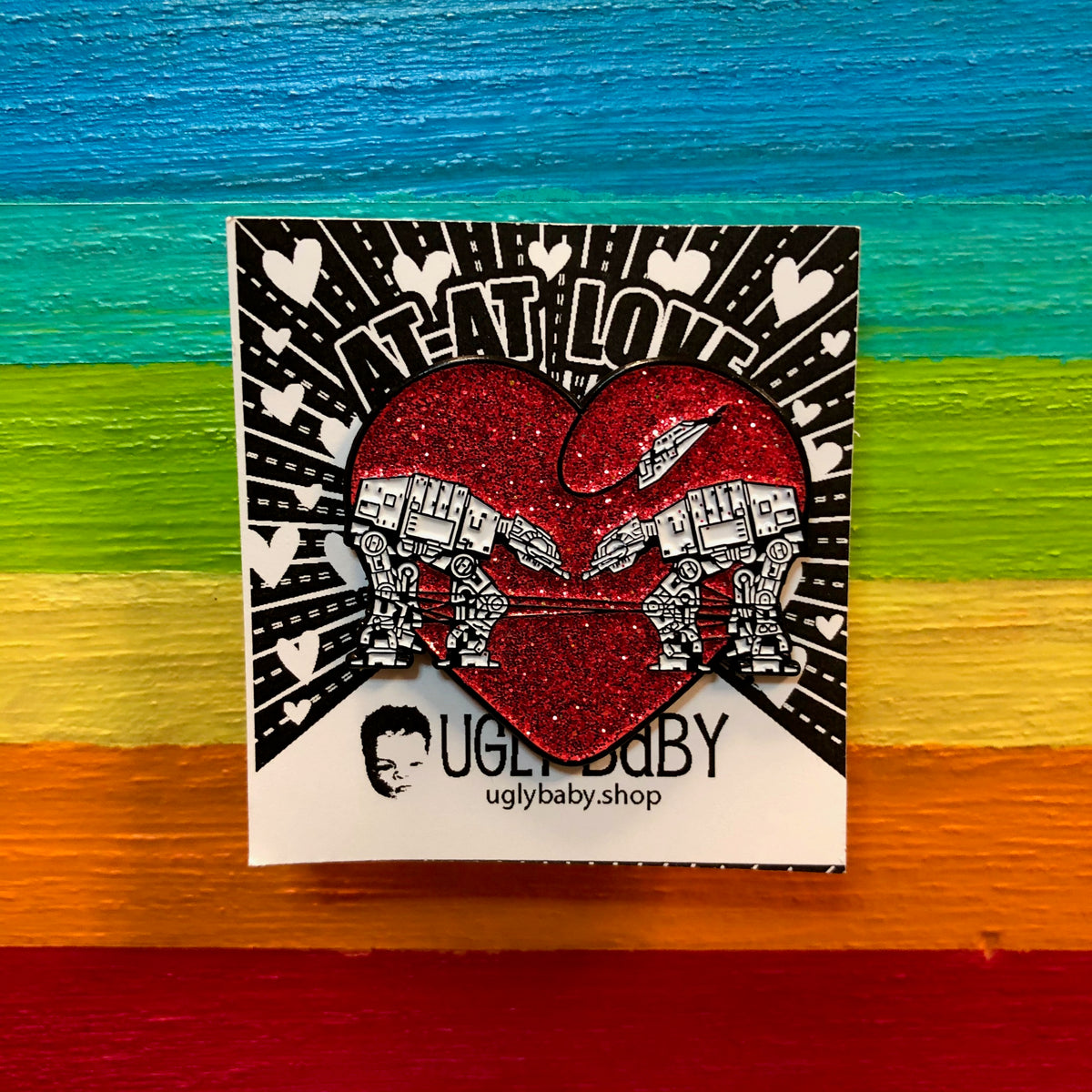 Heart shaped enamel pin called Love AT-AT First Sight with a red glitter background. Shown on a square paper background with black and white hearts. 