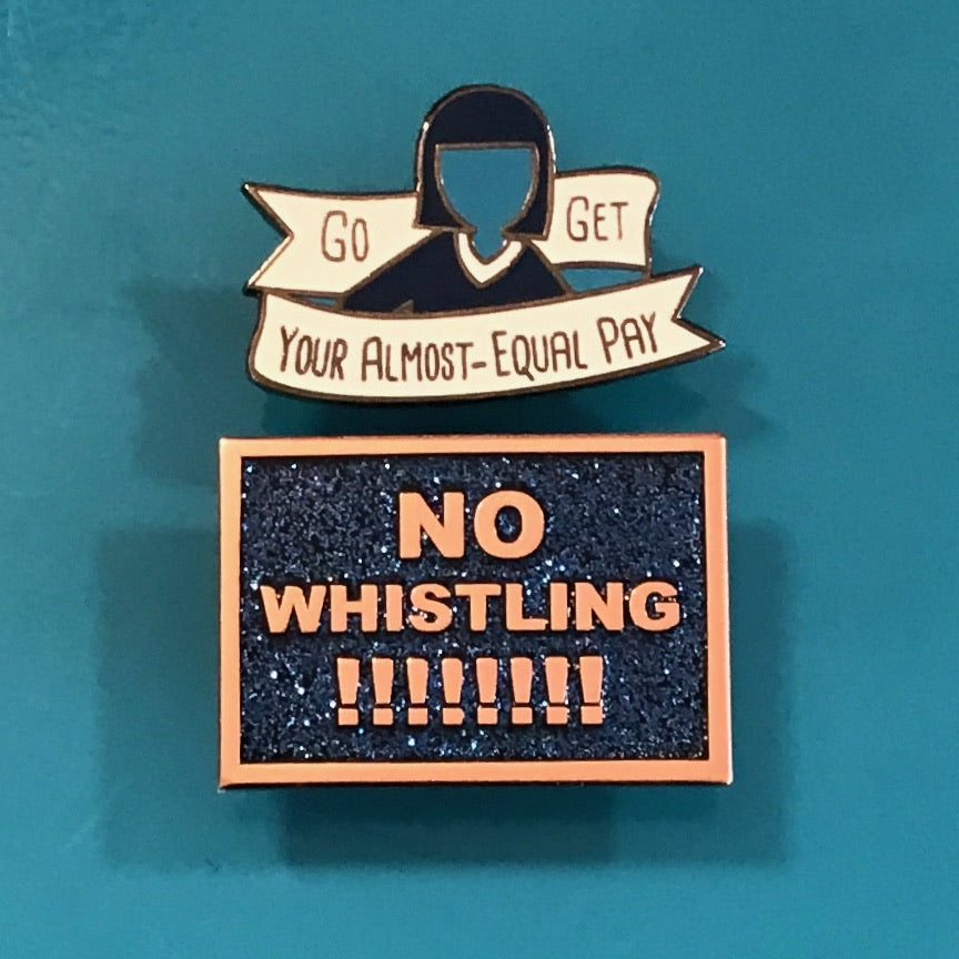 A black with rainbow glitter enamel pin that says "No Whistling !!!!!!!!" in gold text and a blue and white enamel pin that says "Go Get Your Almost Equal Pay." 