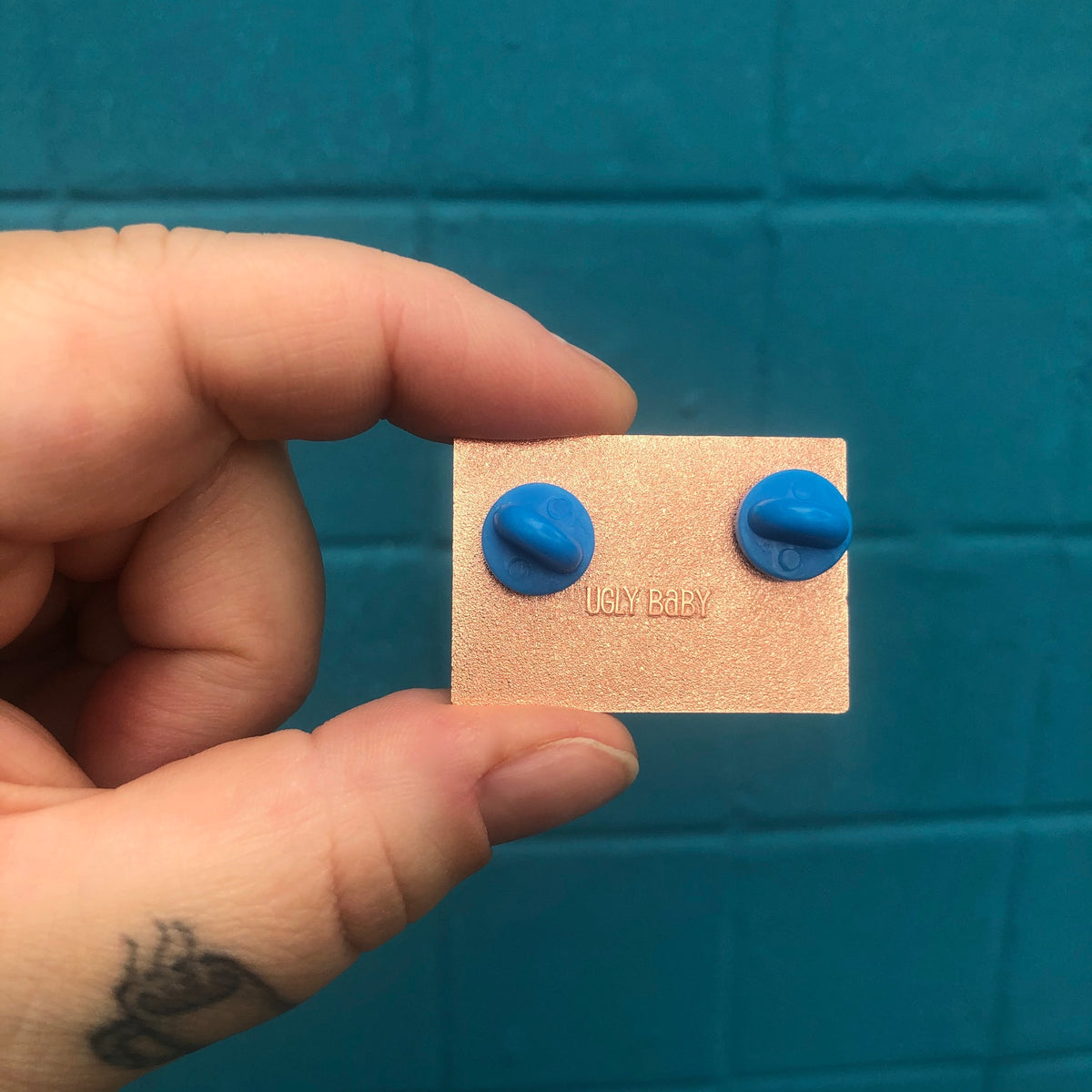 The back of an enamel pin being shown by a human hand against a teal background. The pin has two blue rubber backers and has the words "Ugly Baby" stamped into the back. 