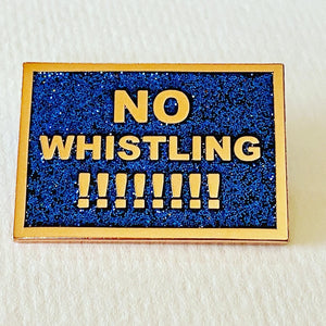 A black with rainbow glitter enamel pin that says "No Whistling !!!!!!!!" in gold text. 