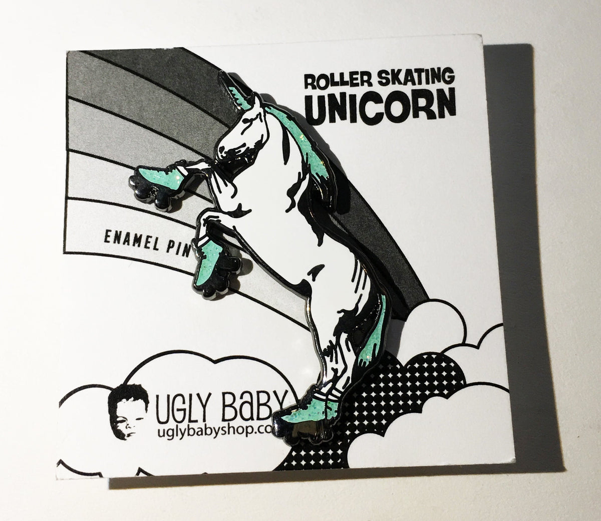 An pin featuring a roller-skating unicorn, mounted on its original white backing card. The backing card displays the text 'ROLLER SKATING UNICORN' and 'ENAMEL PIN' along with the brand 'UGLY BABY' and website 'uglybabyshop.