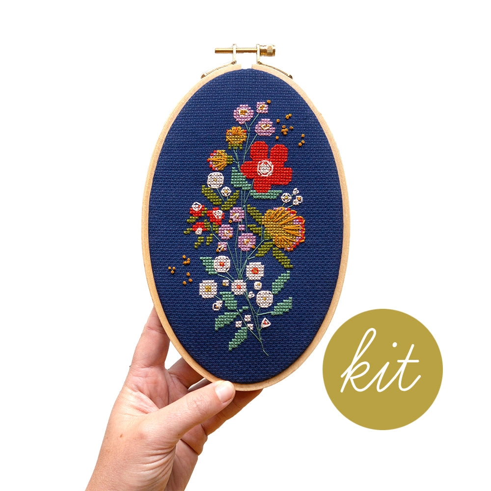 A completed, stitched up example of the Forget Me Knots Cross Stitch Kit is being held up by a human hand. 