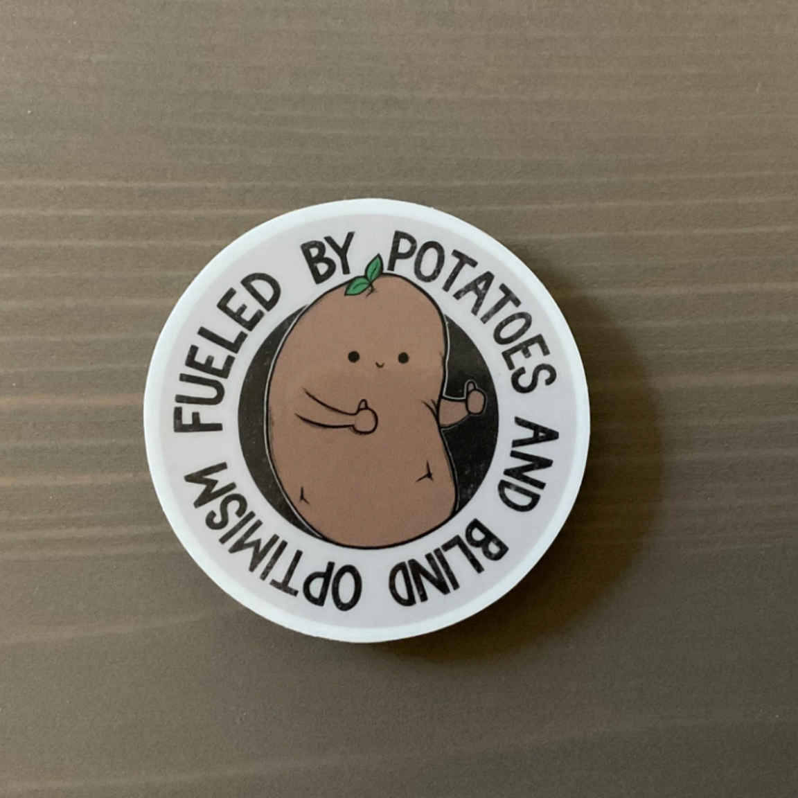 A round sticker that has a drawing of a potato giving two thumbs up and the words "Fueled by potatoes and blind optimism"