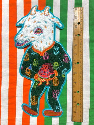 Embroidered patch of a goat in a green outfit on a striped background with a ruler for scale.