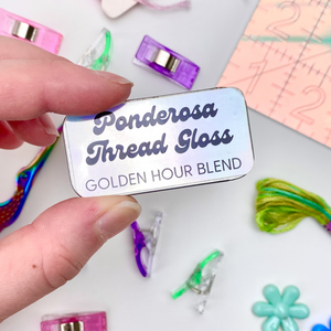 Fingers  holding up a metal tin of Ponderosa Thread Gloss - Golden Hour Blend - against a background of scattered, colorful sewing supplies including: embroidery floss, sewing clips, iridescent scissors, seam ripper and a cutting mat.