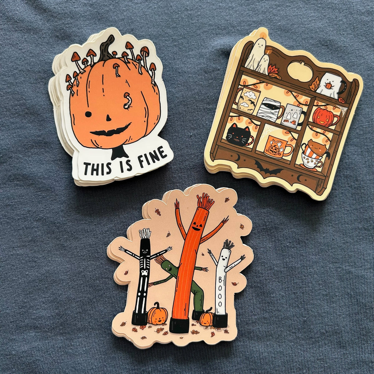 Three Halloween-themed die-cut stickers displayed on a textured slate blue fabric background. The stickers feature whimsical illustrations: a smiling orange pumpkin with mushrooms growing out of its head and the text 'THIS IS FINE', a haunted house shelf scene with cute ghostly characters, and three stick-like figures (black, orange, and white) with arms raised under falling leaves.