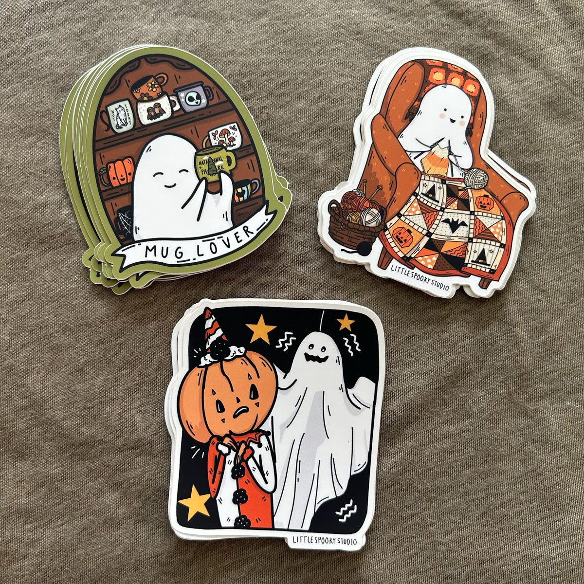 Three die-cut stickers featuring cute cartoon ghost illustrations on a textured taupe fabric background. The stickers include designs labeled 'MUG LOVER', 'KNITTING GHOSTIE', and 'JUMPSCARE'.