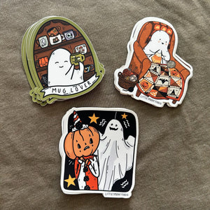 Three die-cut stickers featuring cute cartoon ghost illustrations on a textured taupe fabric background. The stickers include designs labeled 'MUG LOVER', 'KNITTING GHOSTIE', and 'JUMPSCARE'.