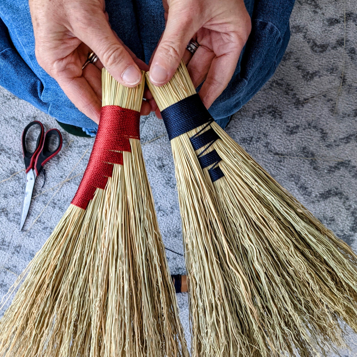 Examples of two hand brooms - one with red detail and another with blue. Two hands are holding them up and you can see a piar of scissors on the floor in the background.
