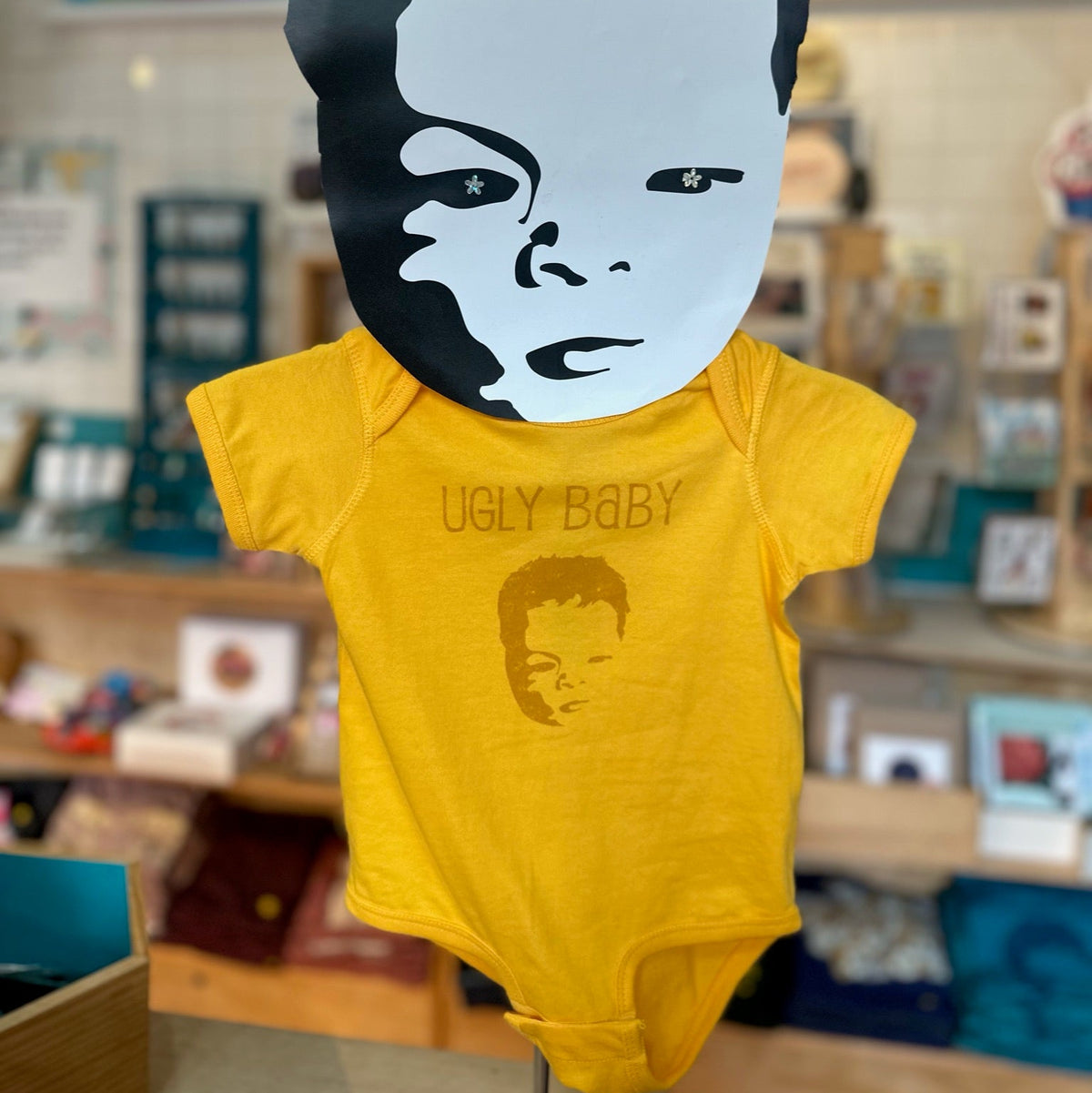 Yellow baby bodysuit with the ugly baby logo on a store shelf background