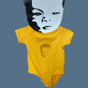 Gold colored infant bodysuit with the Ugly Baby logo printed in darker gold ink. The rack that the bodysuit is hanging on has a large, oversized head in the shape of the Ugly Baby logo at the top. 