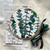 An example of the finished embroidery kit eucalyptus with different shades of green. A cloth with the printed pattern is folded below the example and a batch of embroidery floss is tied with a ribbon. 