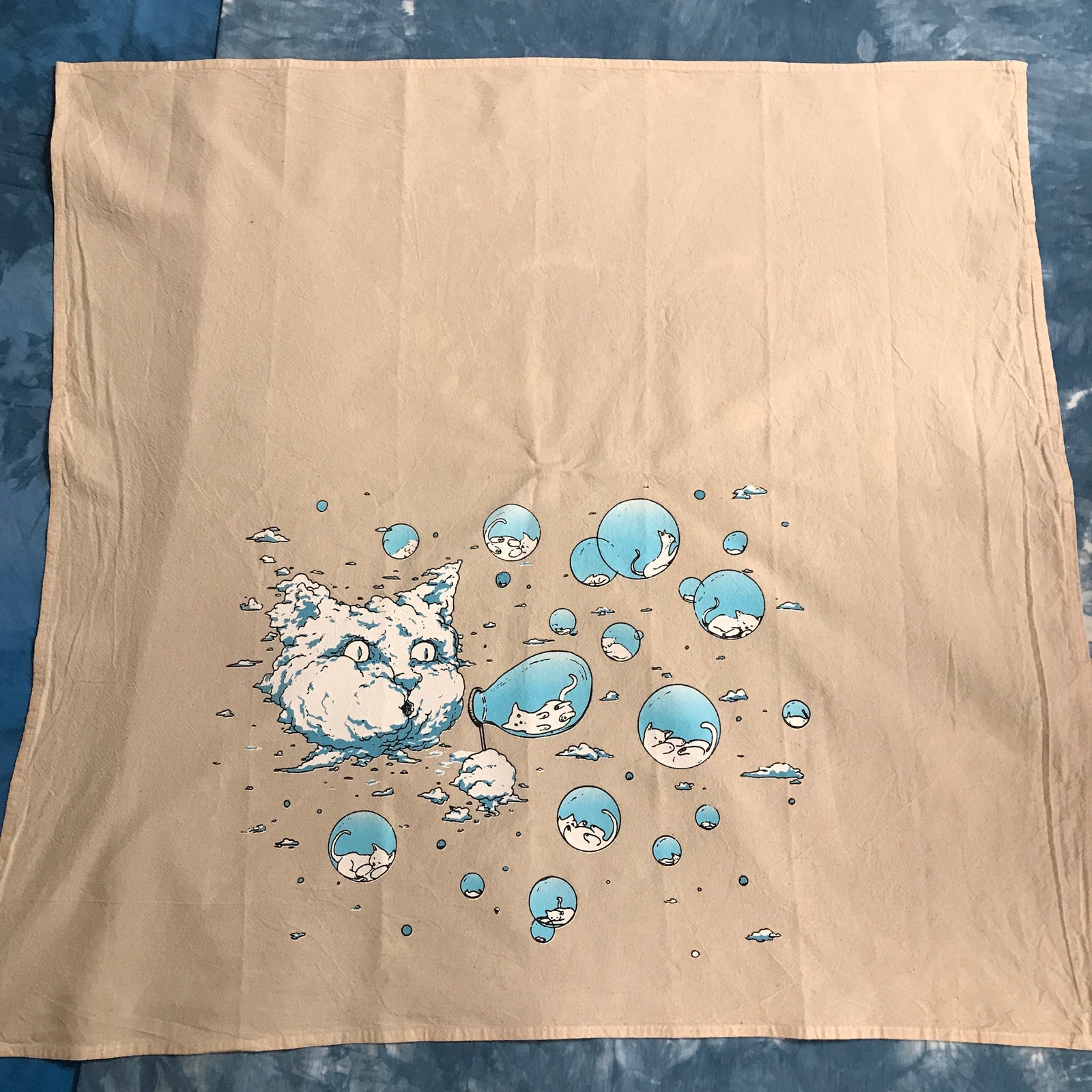A square beige kitchen towel, laid flat on a blue mottled surface. The fabric features a printed graphic of a white cloud cat surrounded by blue and white bubbles. Inside the bubbles are tiny kittens.