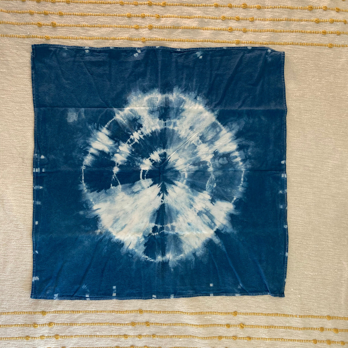 A square blue fabric kitchen towel featuring a white shibori tie-dye spiral pattern, laid flat on a cream-colored embroidered textile background.