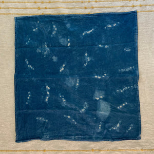 A square indigo blue fabric kitchen towel with white tie-dye shibori patterns, laid flat on a cream-colored embroidered textile background.