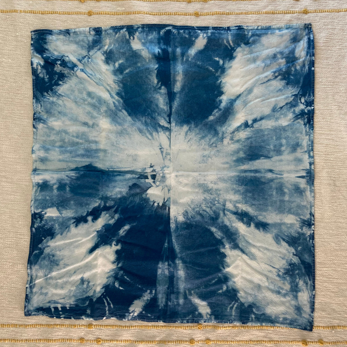 A square fabric kitchen towel with a blue and white shibori tie-dye burst pattern, placed on a cream-colored textured textile with gold embroidered horizontal stripes.