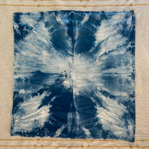 A square fabric kitchen towel with a blue and white shibori tie-dye burst pattern, placed on a cream-colored textured textile with gold embroidered horizontal stripes.