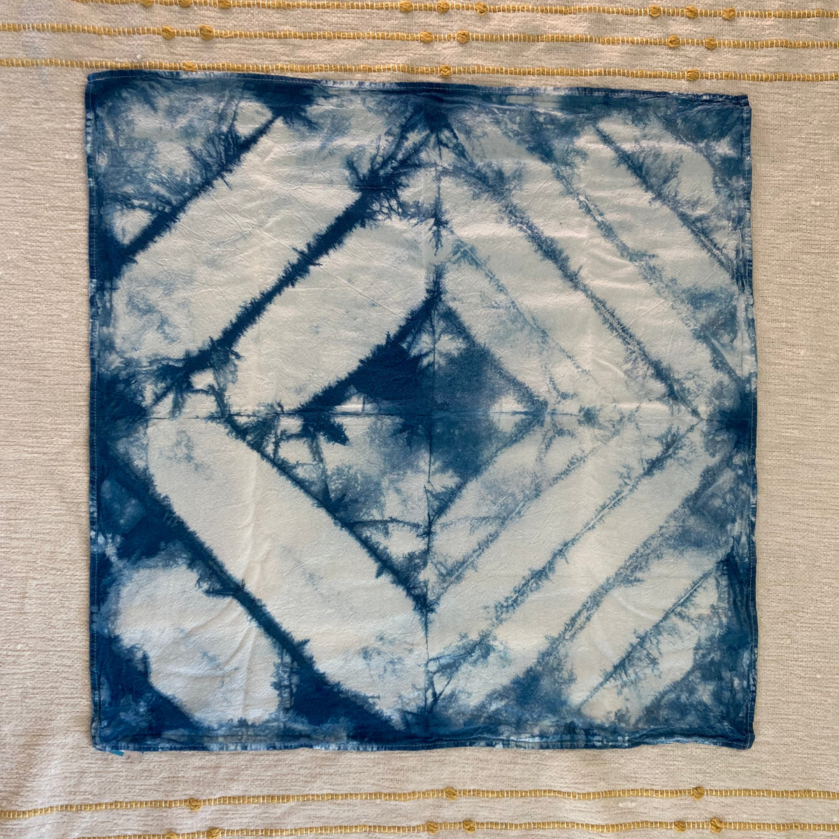 A square fabric kitchen towel with a blue and white tie-dye geometric pattern, laid flat on a textured cream-colored fabric background.