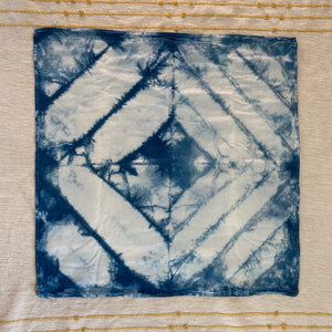 A square fabric kitchen towel with a blue and white tie-dye geometric pattern, laid flat on a textured cream-colored fabric background.