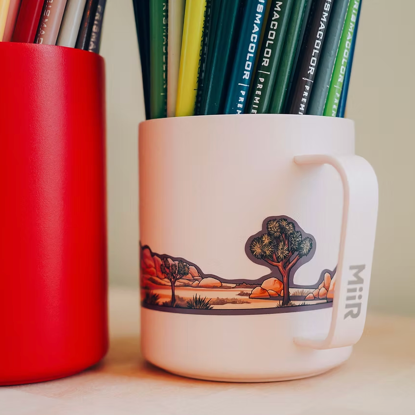 White mug with Joshua Tree National Park design filled with colored pencils, next to a red mug.
