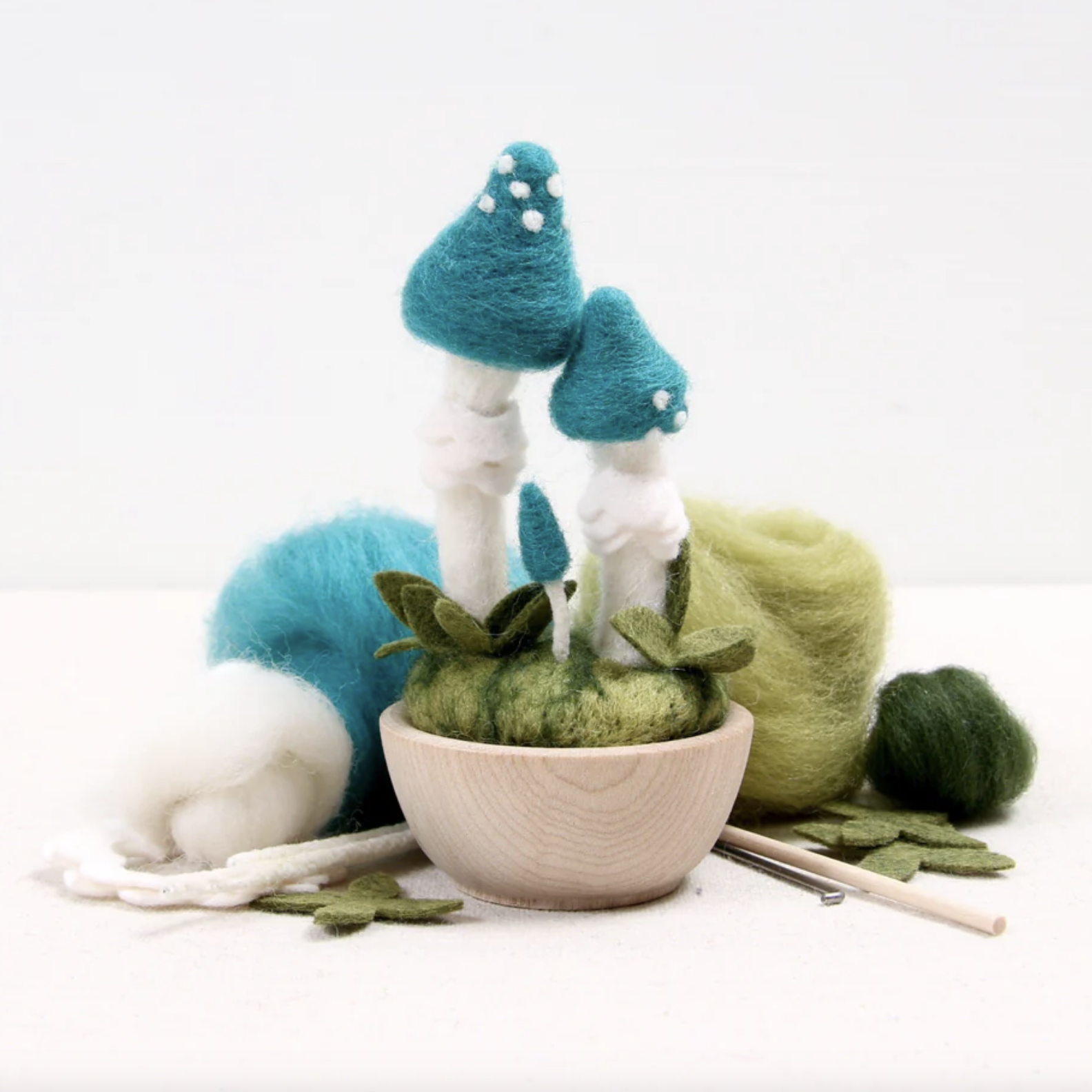 Completed needle felting project of a Blue Roundhead Mushroom. It is shown with needle felting supplies included in the craft kit on the table around it. 