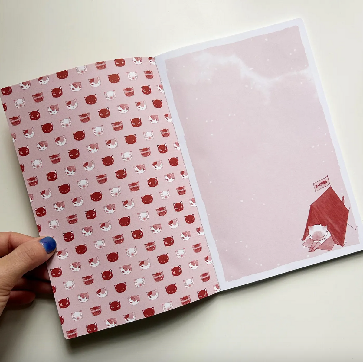 Open notebook with red and white pattern on one page and pink cover with design on the other, held by a hand.