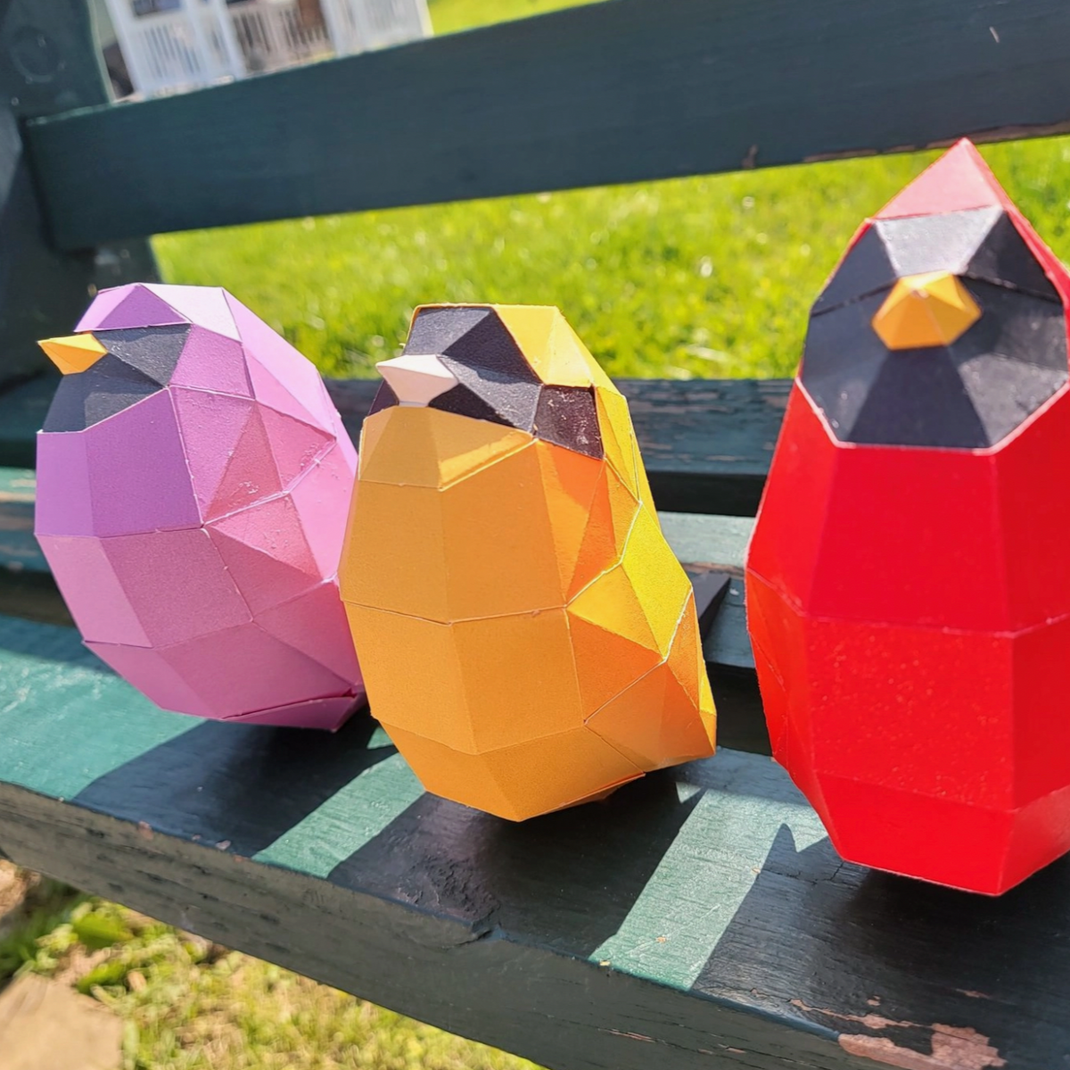 Examples of paper craft bird kit. They are pink, yellow and red & are all different sizes. They are sitting on a park bench with grass in the background.