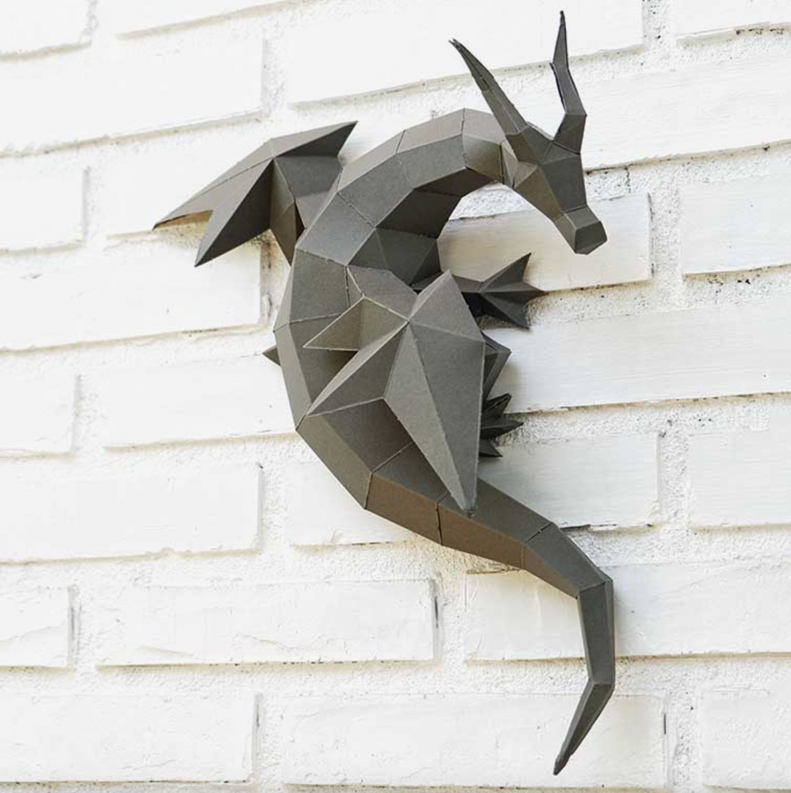 An example of a dragon paper craft kit completed and hanging on a white brick wall.