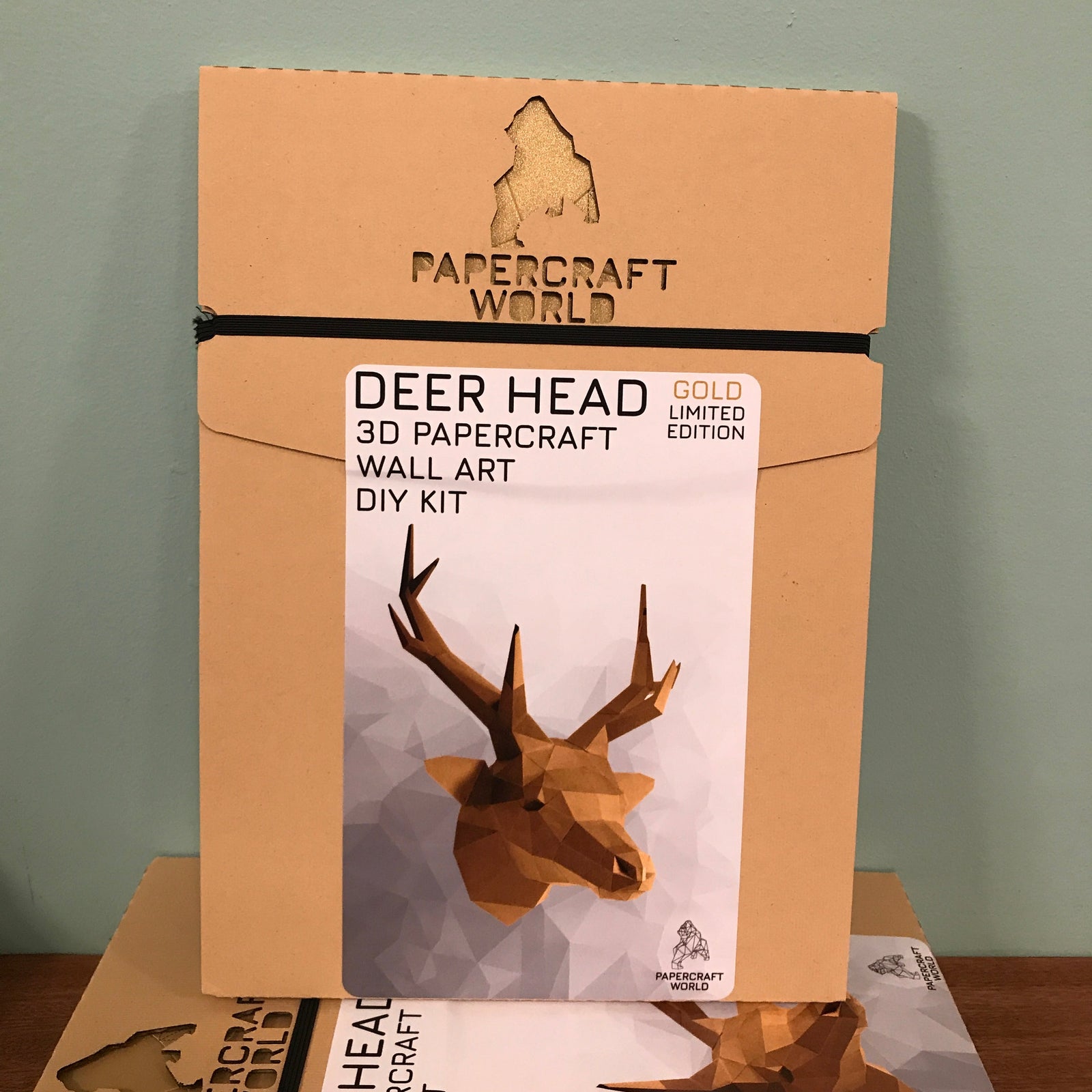 Packaging for the gold deer hear. It is kraft colored and has the papercraft world logo laser cut into the top. It has a picture of a completed gold deer head on the front.
