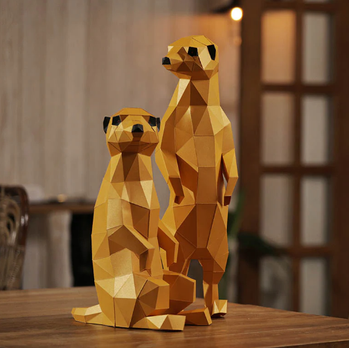 Completed examples of the meerkats in the paper craft kit. They are two different sizes and are standing on a table with a dining room in the background.