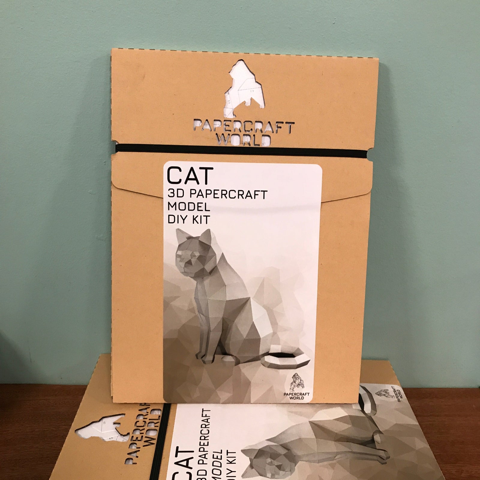 Packaging for the white cat paper craft kit. The package is kraft colored and has the papercraft world logo laser cut into the top. It has an example of the completed white cat paper craft kit.